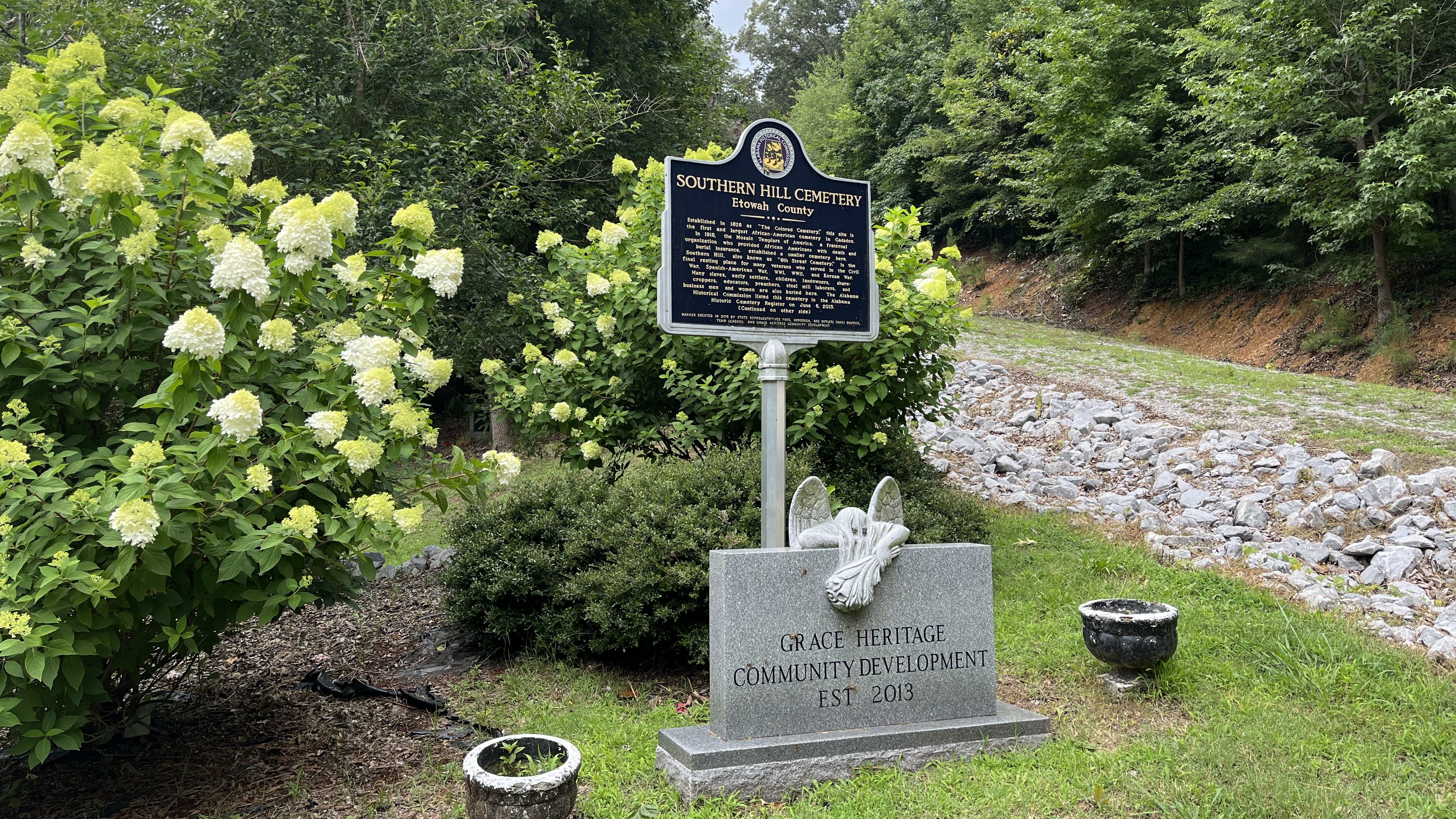 A view of Southern Hill Cemetery with a historic marker and a granite memorial stone in front of flowering bushes and trees.